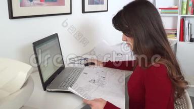 设计工程师坐在她办公室的办公桌前，在笔记本电脑上打字，在她的办公桌上放蓝图